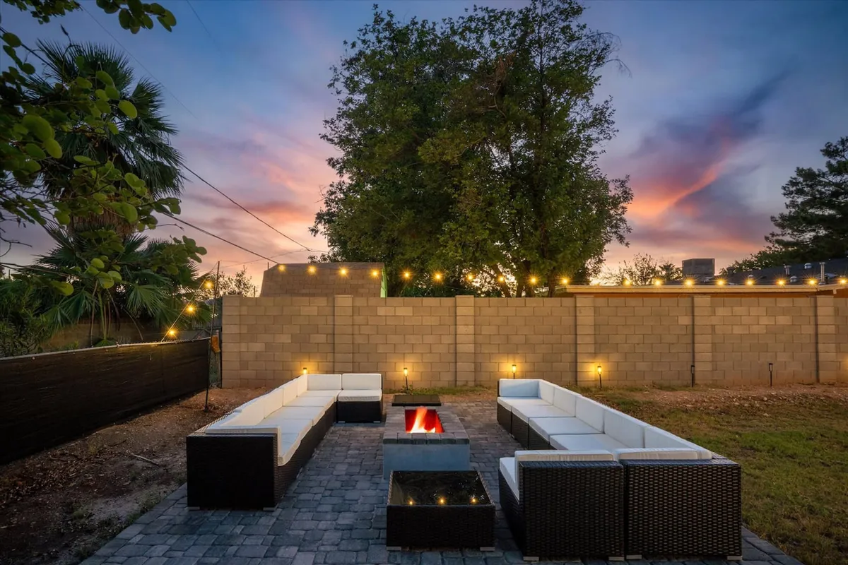 The image showcases a beautifully lit backyard at dusk, centered around a glowing fire pit surrounded by modern outdoor seating. Decorative string lights twinkle overhead, casting a warm ambiance on the lush greenery that frames the scene. The setting is inviting for both relaxing evenings and vibrant social gatherings, with plenty of space for guests to unwind and connect.