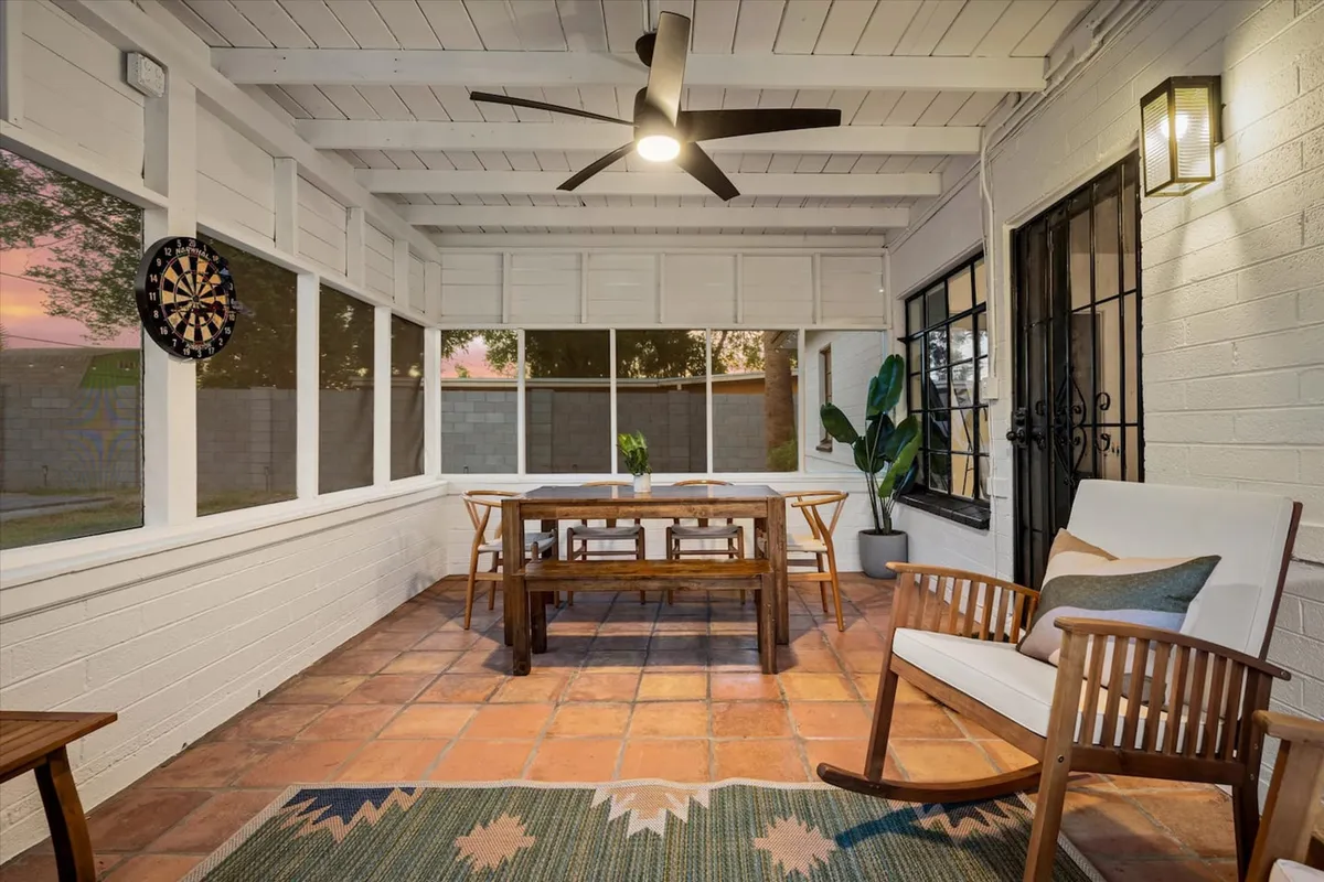 The image features a wooden dining table with a casual setup, a rocking chair nearby, and a dartboard on the wall, all set in a space bathed in warm, inviting light. Potted plants add a touch of greenery, and the overall vibe is casual and family-friendly.