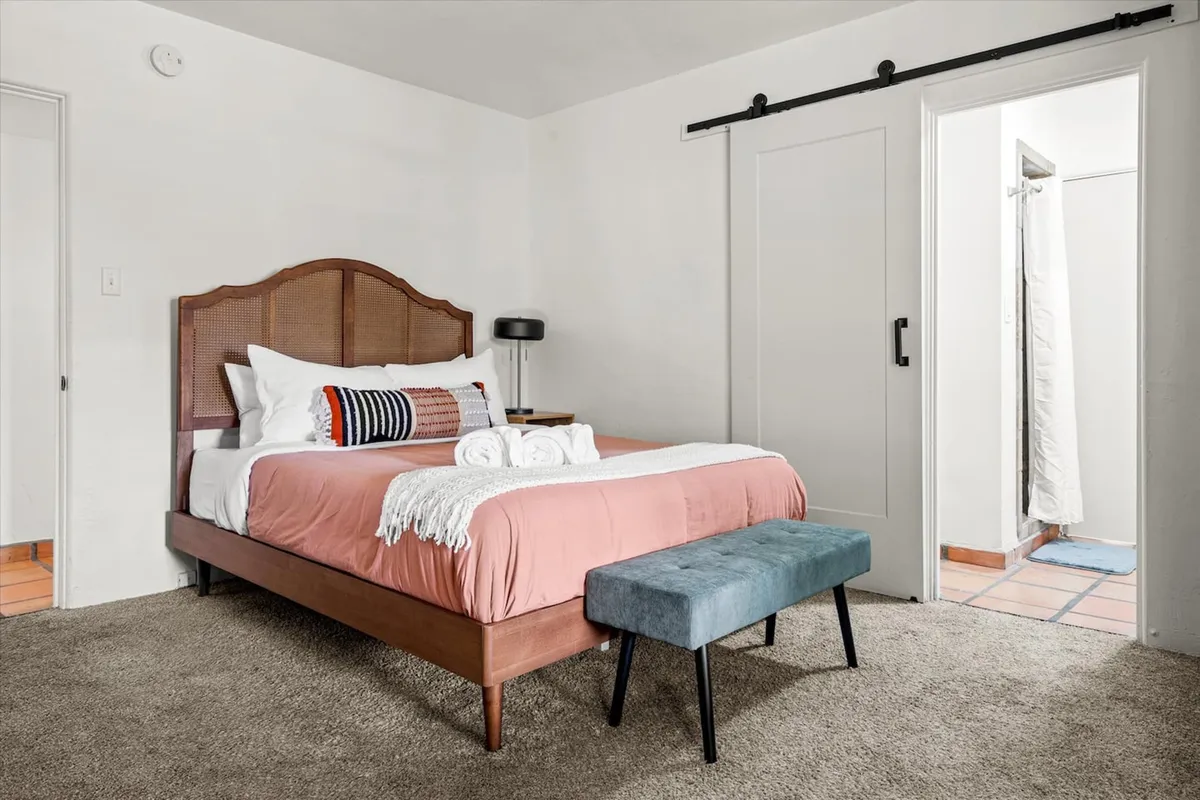 A calm bedroom scene with a plush bed, decorative pillows, a welcoming bench at the foot of the bed, and a visible doorway leading to a bathroom. The soft lighting and subtle design details suggest a seamless flow between private sleeping and bathing spaces.