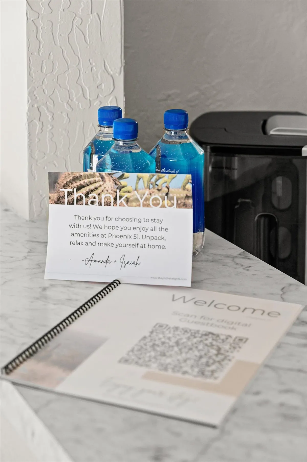 A close-up of a welcome vignette featuring a thank you note, several water bottles, and a welcome booklet arranged on a surface. The mood is friendly and inviting, suggesting attentive, concierge-style guest services.