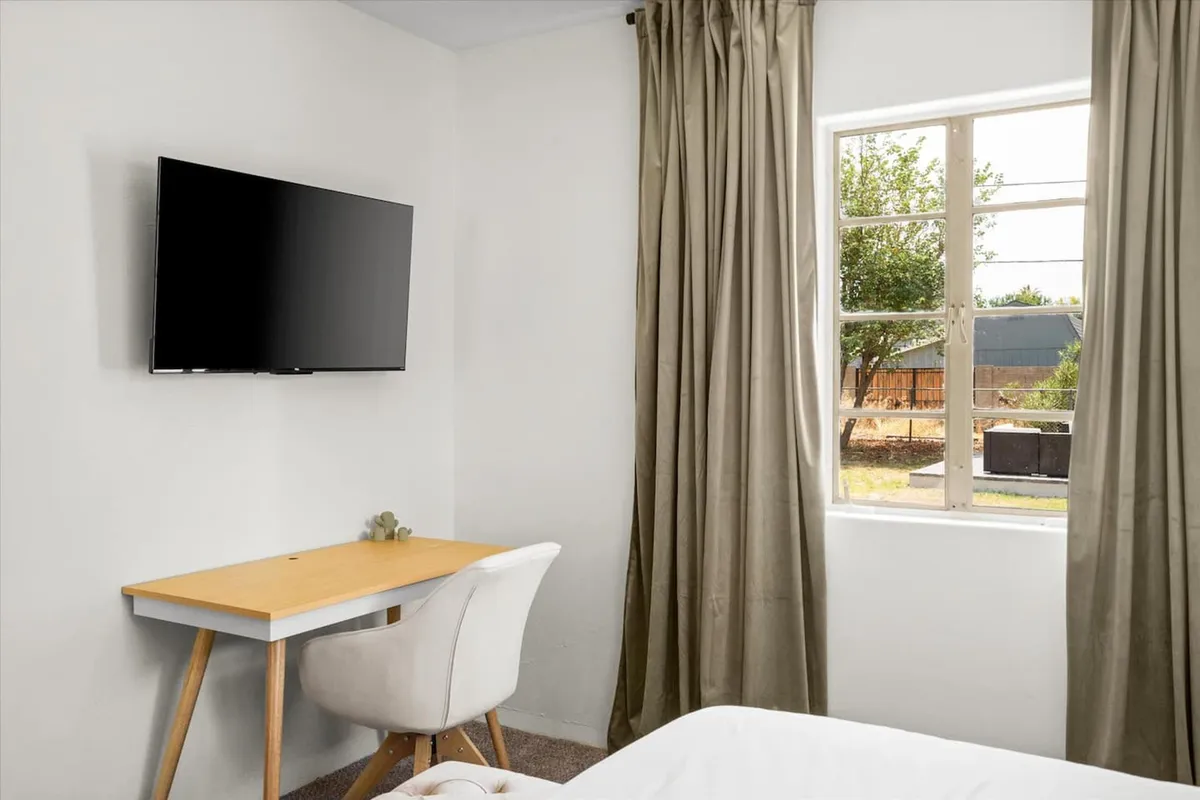 A cozy workspace featuring a television, a modern desk and chair, and a window allowing in natural light. The setup is ideal for both productivity and relaxation, with space to work, watch TV, or enjoy quiet moments.