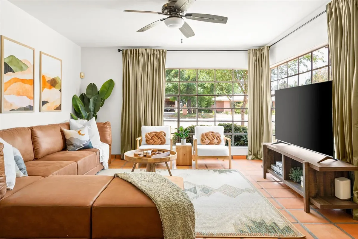 A spacious living room featuring a comfortable brown sectional sofa positioned to face a modern TV on a sleek stand. Large windows allow natural light to flood the space, highlighting decorative wall art and a coffee table adorned with tasteful decor. Lush greenery is visible through the windows, lending a fresh, inviting feel.