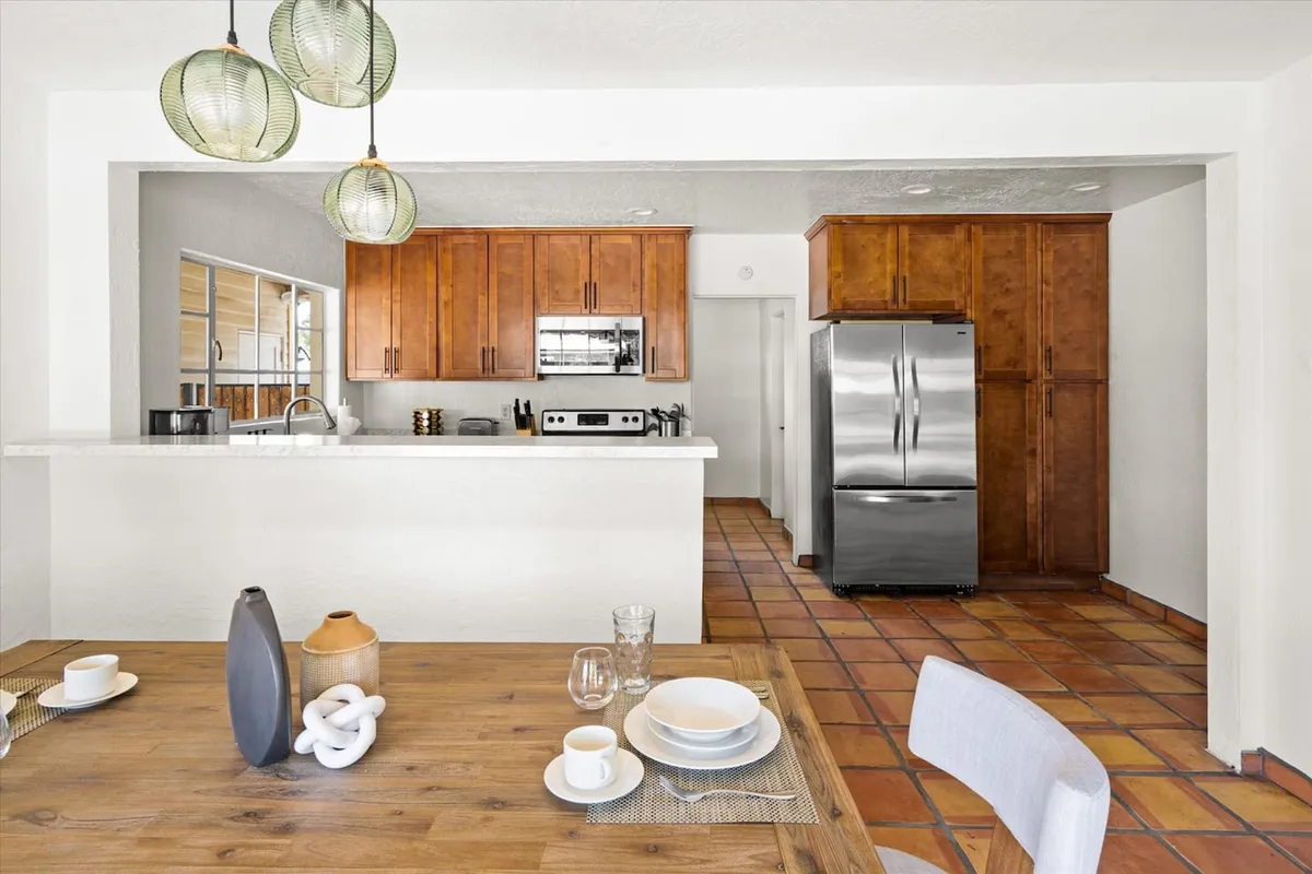 A spacious, modern kitchen and dining area featuring a kitchen island with pendant lighting, a stainless steel refrigerator, and warm wood cabinetry. The open layout invites group cooking and socializing, with ample space for meal prep and gathering.