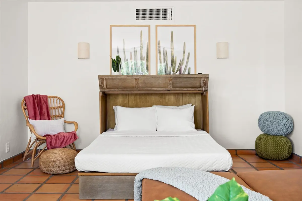 A serene bedroom featuring a bed dressed in crisp white linens, a wooden headboard, cactus-themed artwork, and a rattan chair. Natural light filters softly into the room, creating an inviting, tranquil atmosphere ideal for relaxation and restful sleep.