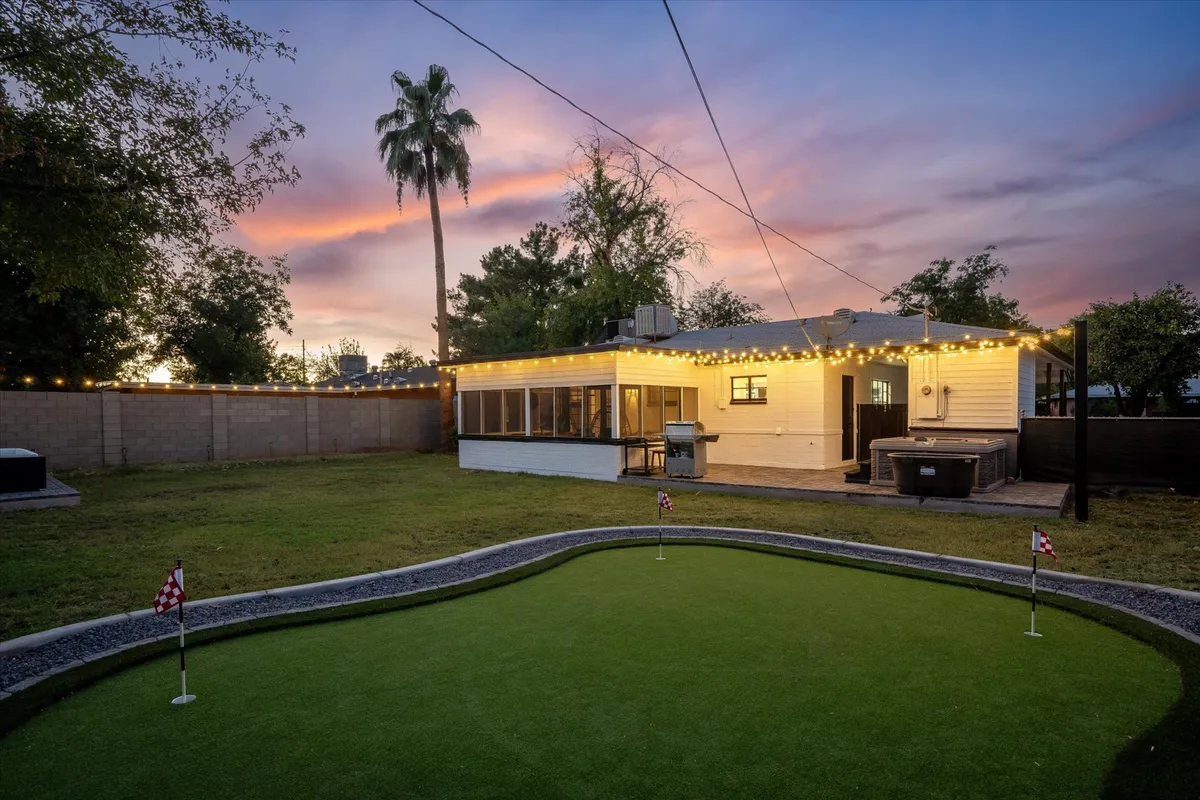 A sweeping view of the backyard featuring a lush putting green bordered by palm trees, an inviting outdoor seating area beneath a canopy of string lights, and the edge of a hot tub visible on the side. The setting is sunlit, evoking a cheerful, relaxed atmosphere ideal for both entertaining and unwinding.