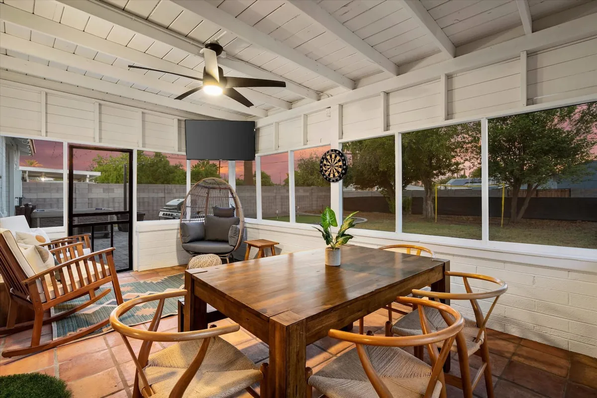 A cozy dining area with a wooden table, a hanging chair nearby, large windows letting in natural light, and decor including a dartboard and indoor plants. The scene suggests a relaxed, multi-use space perfect for meals, games, or coffee breaks.