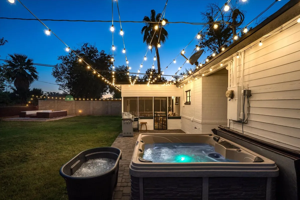 A cozy, resort-style backyard scene at dusk features a shimmering hot tub illuminated from within, with comfortable outdoor seating and overhead string lights casting a warm, inviting glow. The setting feels both tranquil and festive, ideal for soaking under the stars or entertaining guests.