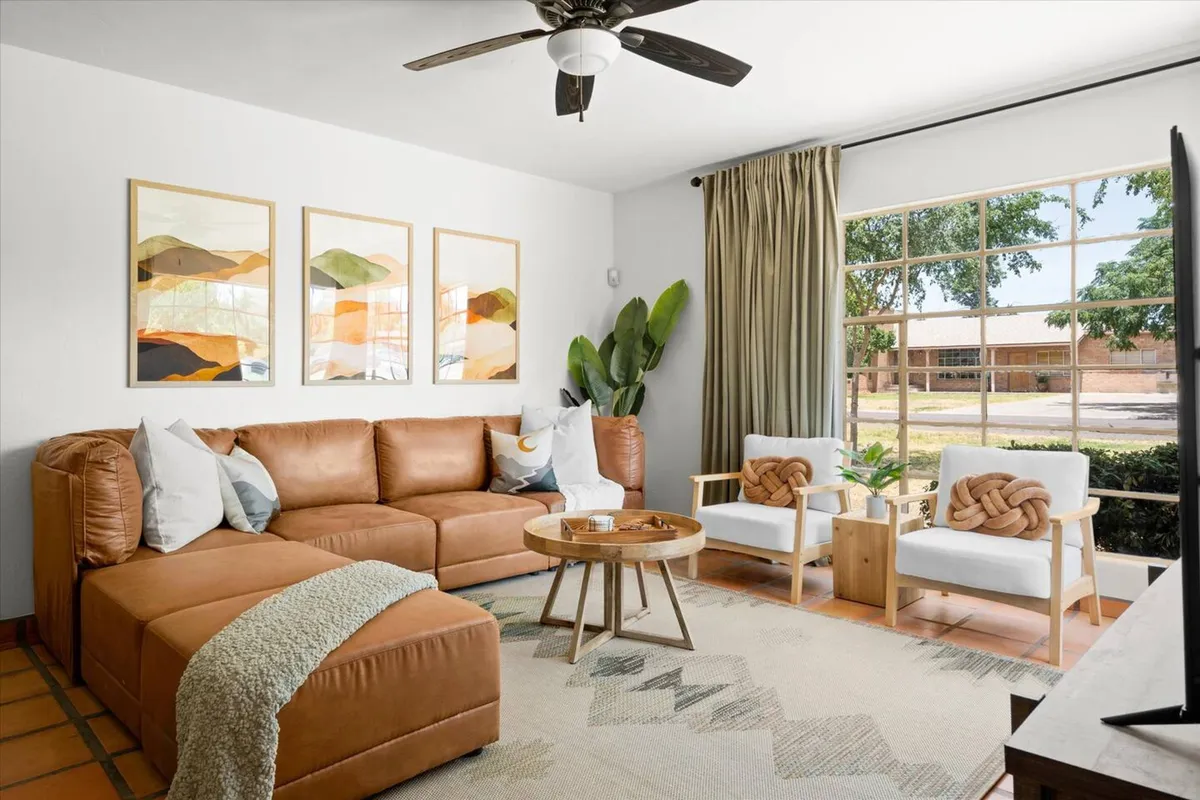 A contemporary living area with a plush leather sectional sofa, colorful wall art, a large window showcasing greenery, wooden accent chairs, and a round coffee table. The space feels welcoming and serene.