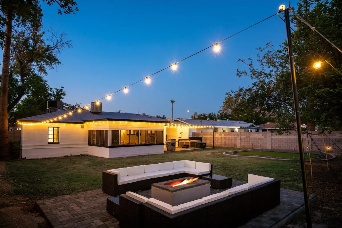 This exterior image features a stylish backyard setup with a fire pit, outdoor seating, string lights, and a lush lawn in front of the residence. The scene evokes a sense of relaxation and togetherness, perfect for unwinding after a day out. The inviting atmosphere suggests opportunities for both relaxation and outdoor activities, aligning with the wellness and self-care theme.